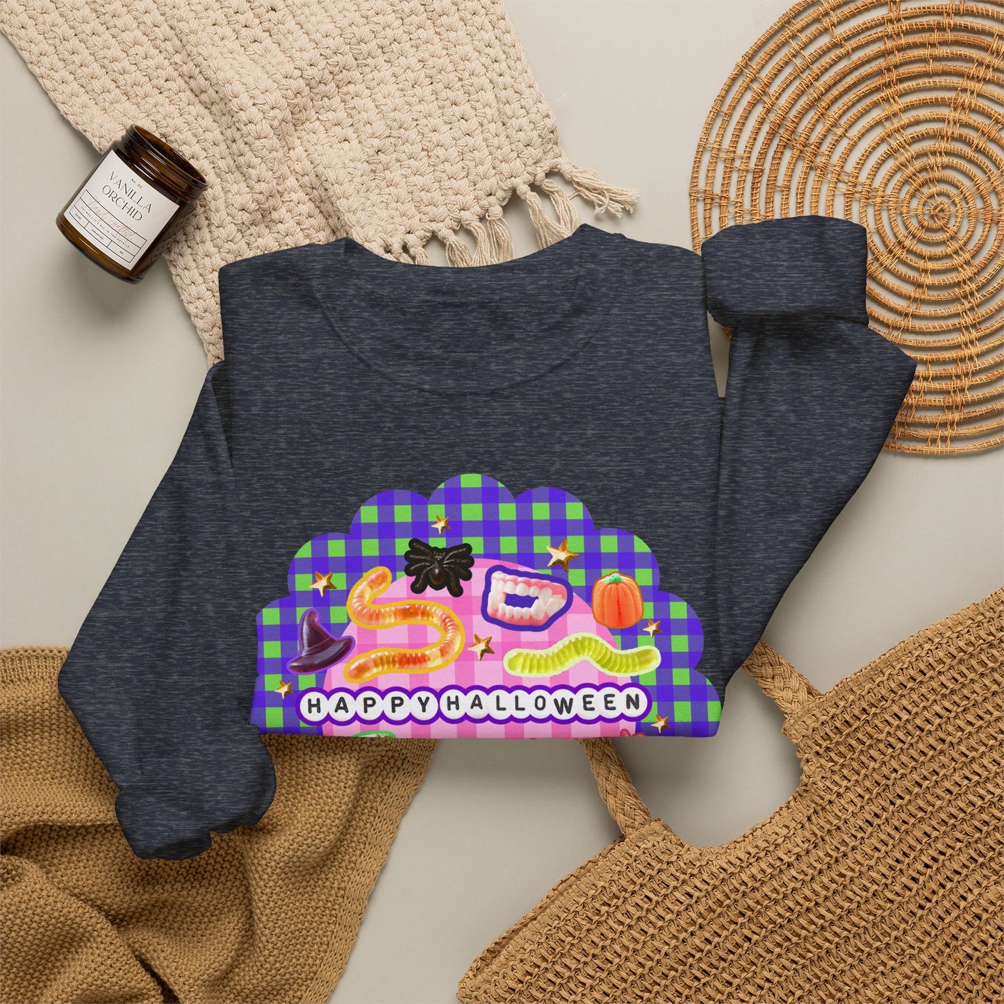 A dark heather gray sweatshirt with a colorful 'Happy Halloween' graphic print, styled in a flat lay with a beige knit blanket, a small candle, and a woven placemat.