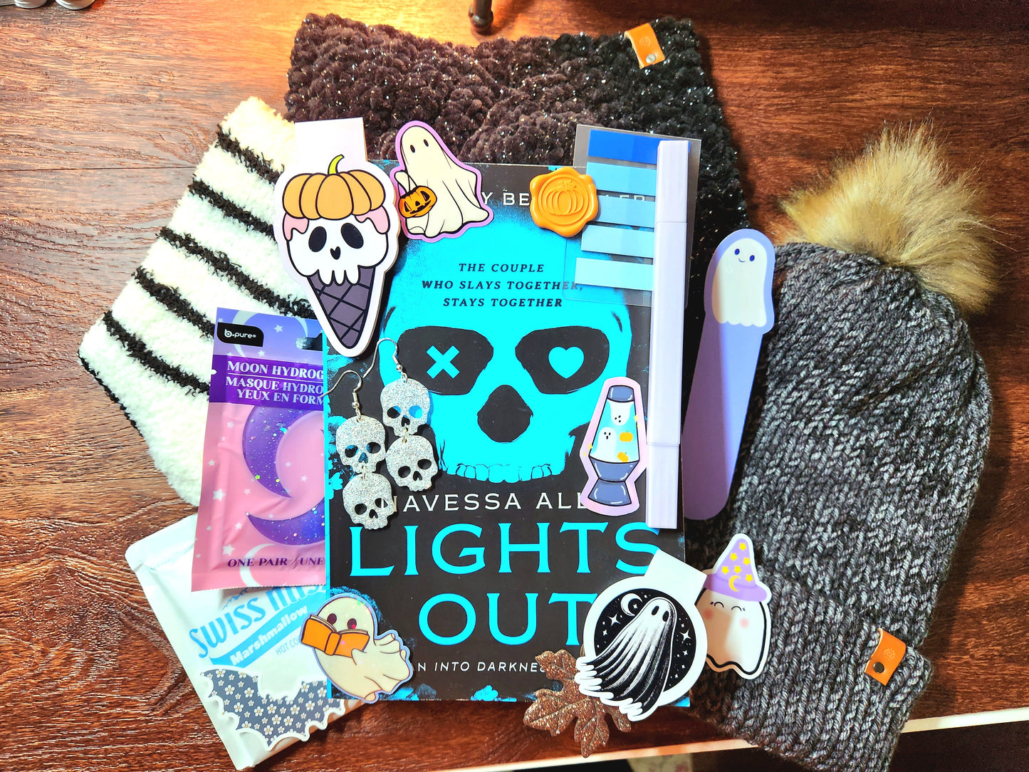 A collection of various items including a book, a hot chocolate, magnetic bookmarks, a headband, a beanie, socks, a highlighter, stickers, earrings, a face mask, and a bookmark, all arranged on a wooden surface.