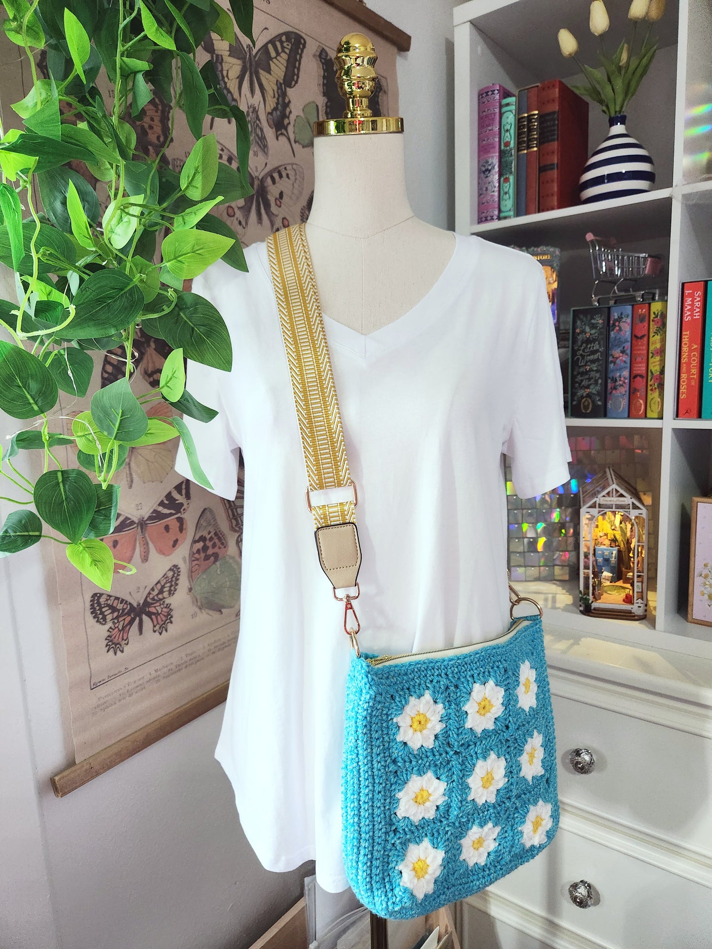 Mannequin wearing a white shirt with a blue crochet bag featuring white flowers, in a room with books and plants.