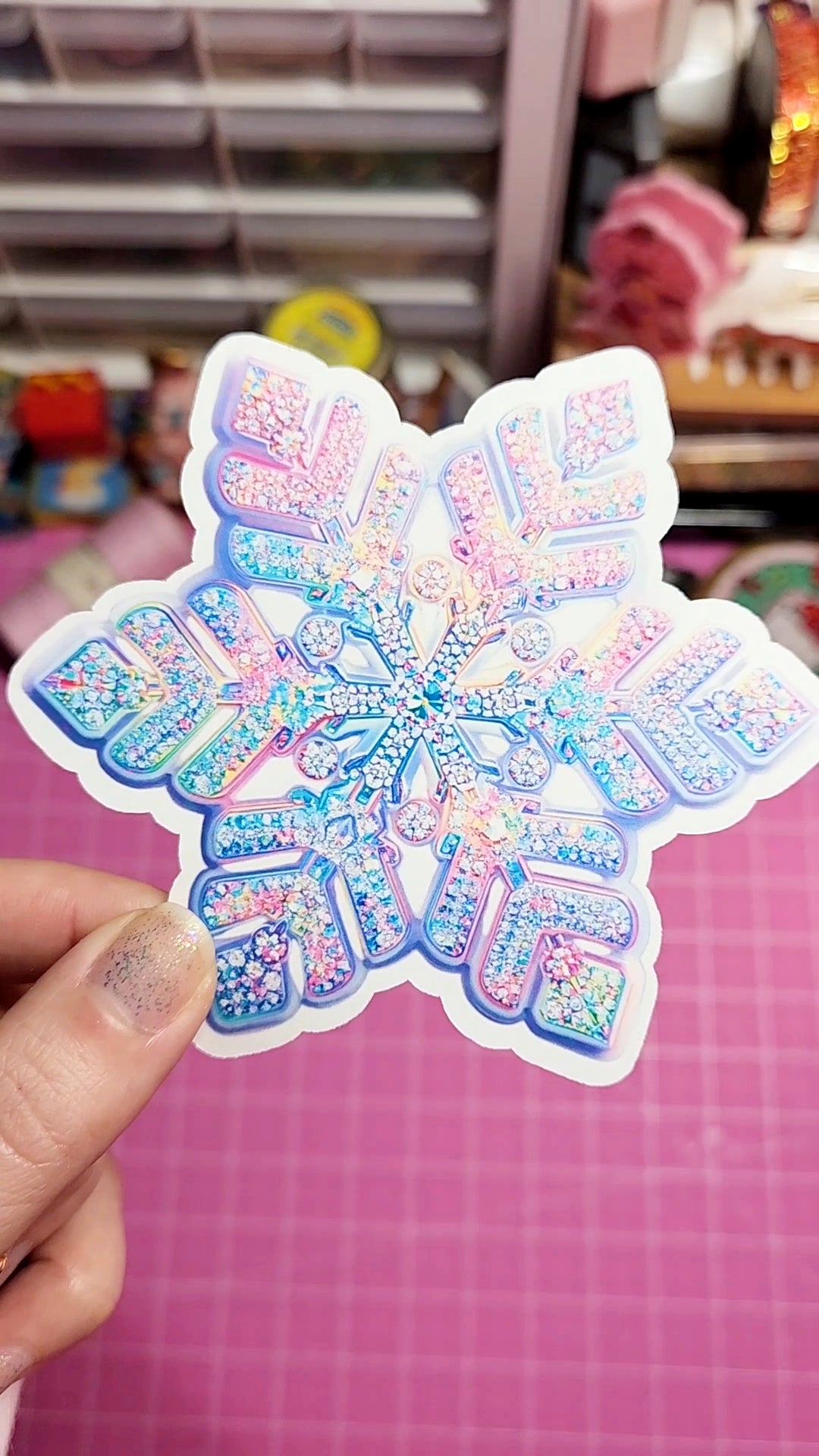 Holographic snowflake-shaped sticker held by a hand on a pink background