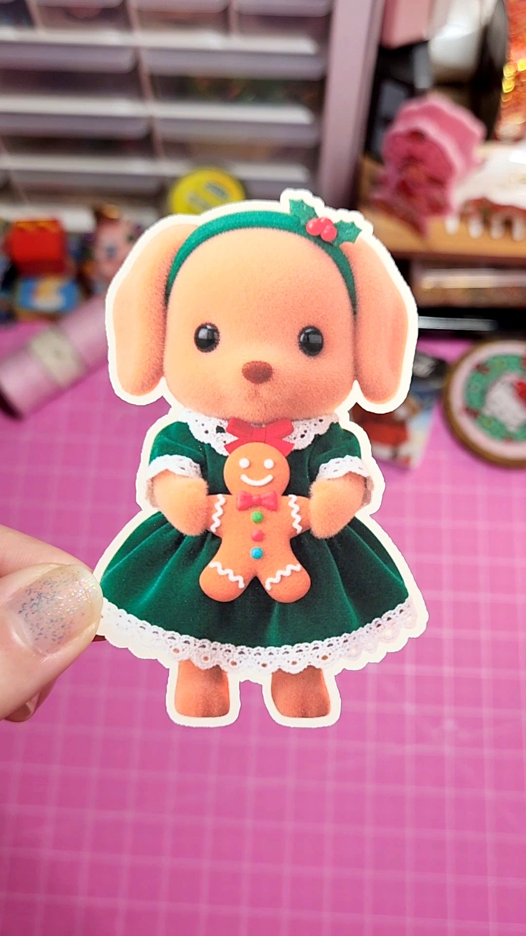 Cartoon dog sticker in a green dress holding a gingerbread man on a pink grid background.