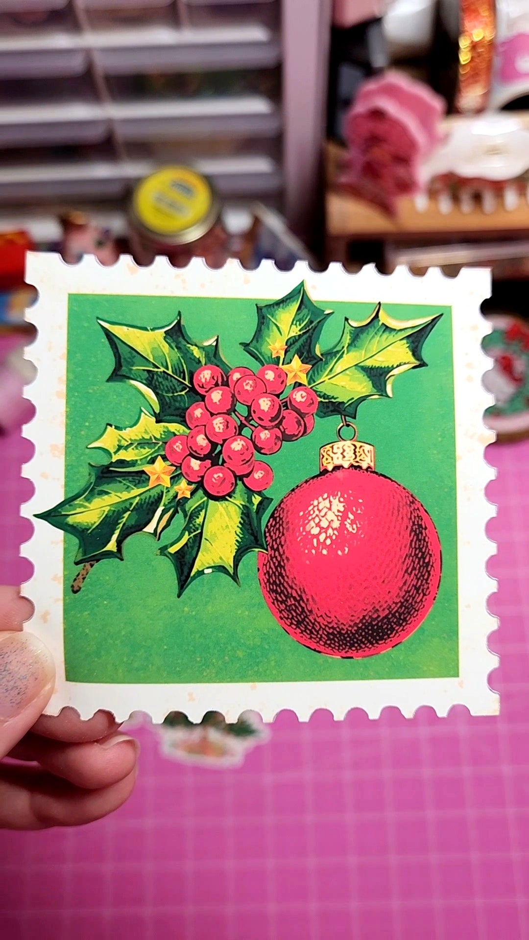 Decorative card with holly leaves and berries and a pink ornament on a green background.
