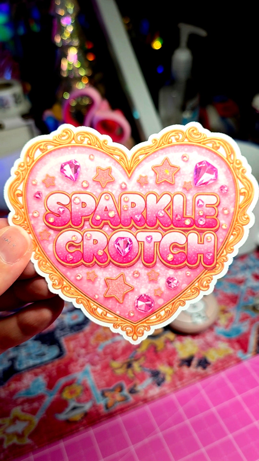 Heart-shaped sticker with 'Sparkle Crotch' text held by a hand on a colorful background.