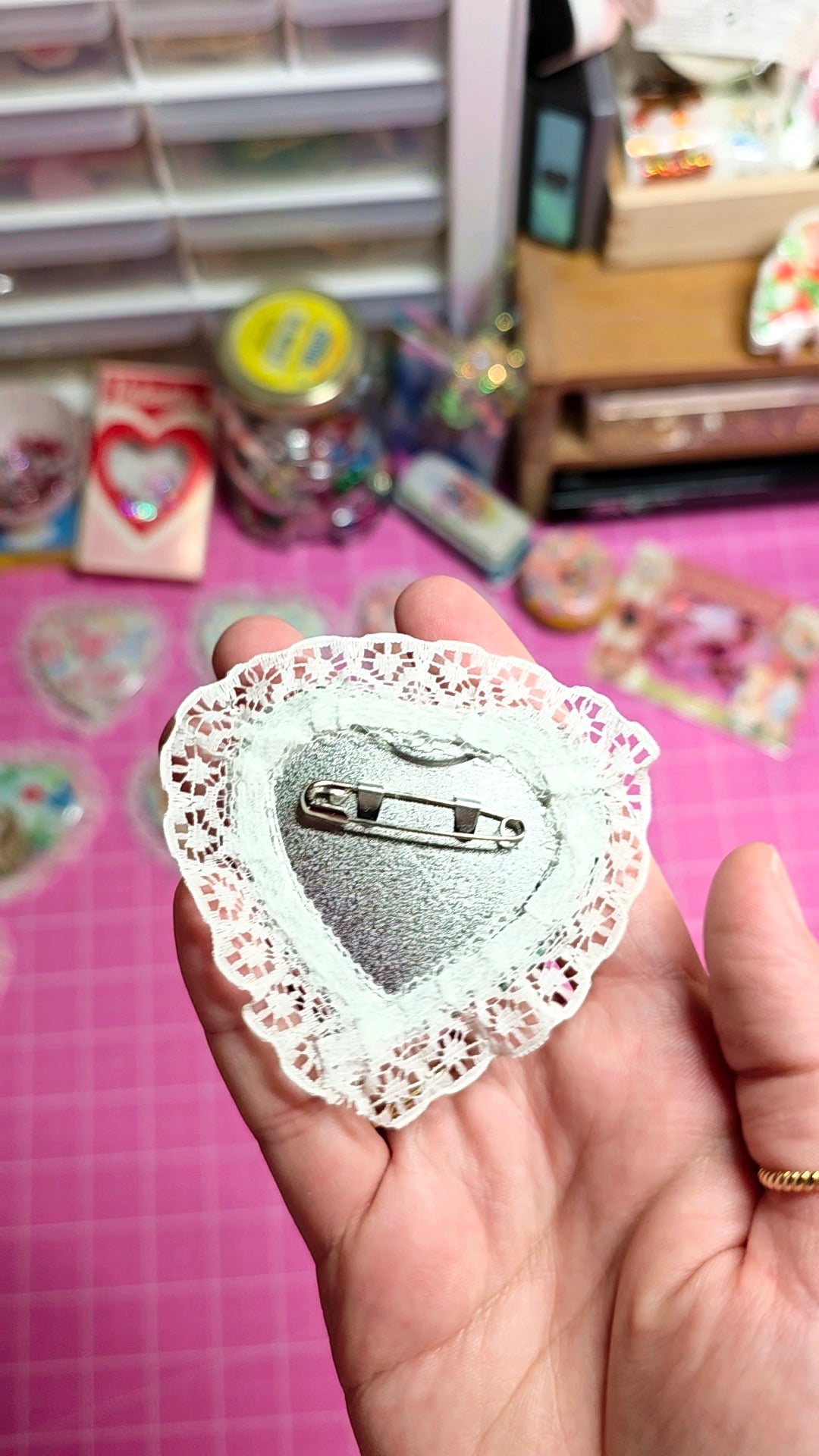 Heart-shaped brooch with lace detailing held in a hand on a pink surface with craft supplies.