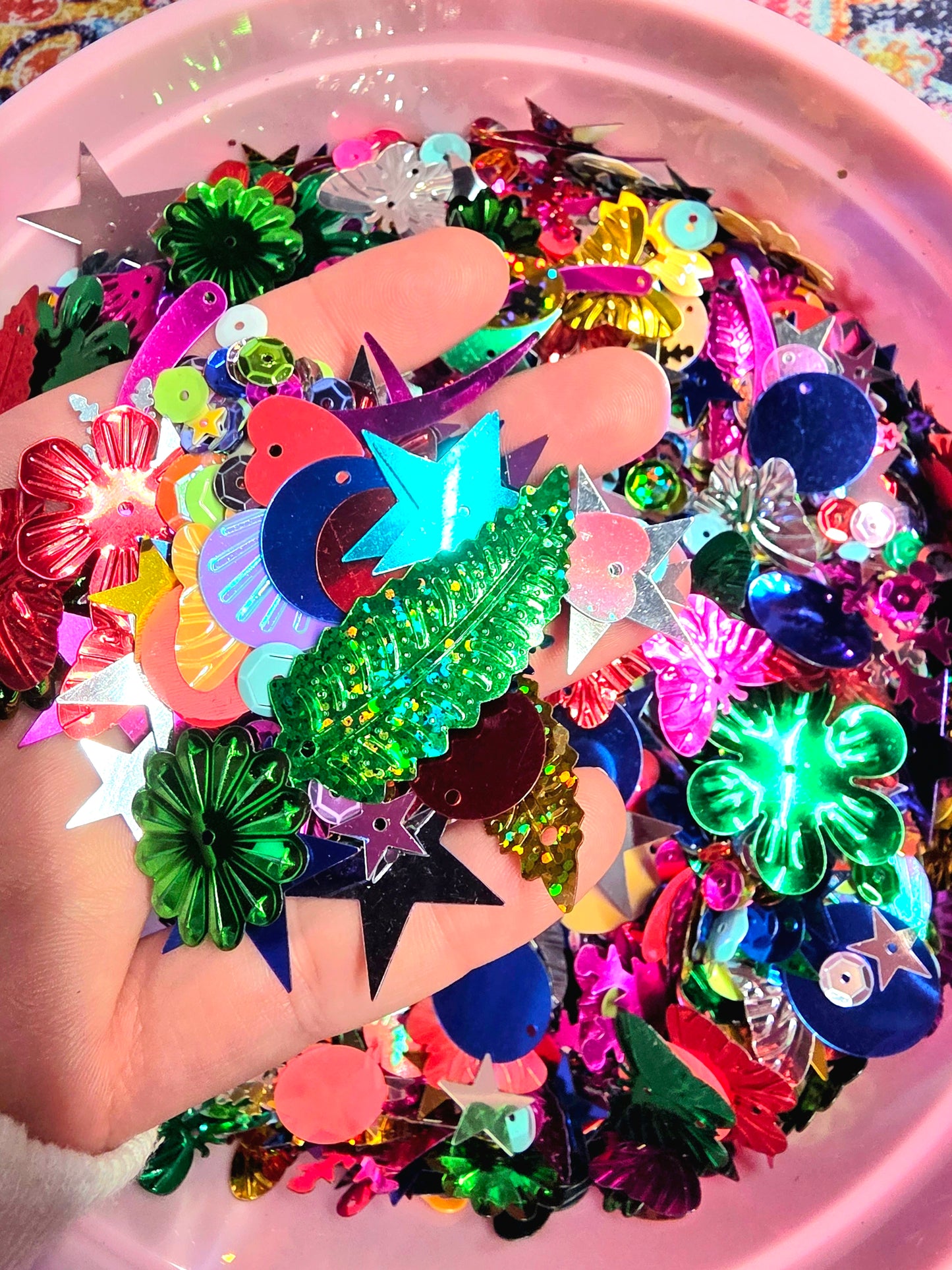 Colorful plastic embellishments held by a hand with a pink container filled with more embellishments in the background.