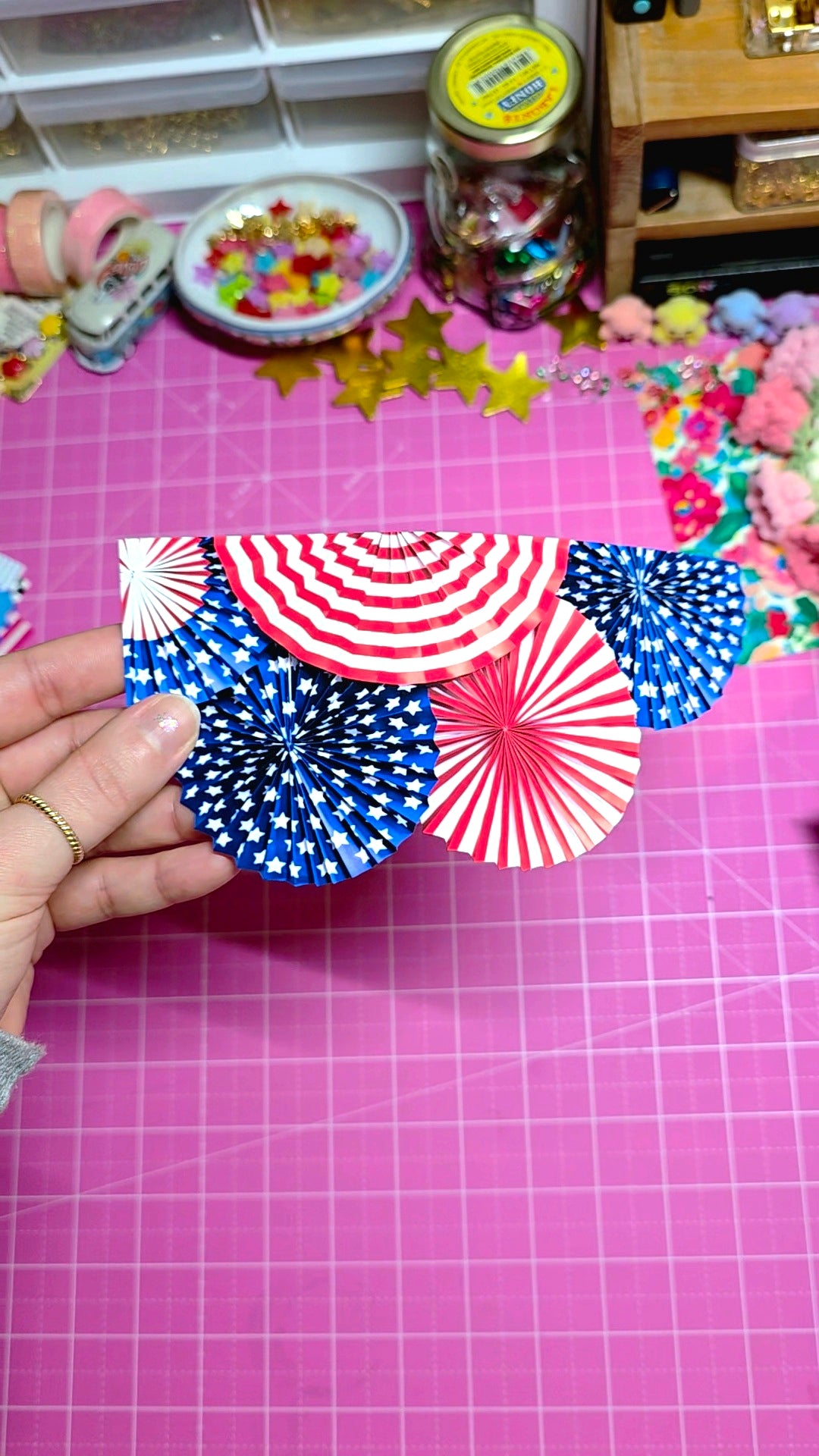 Hand holding a small box with red, white, and blue paper fans on a pink cutting mat.