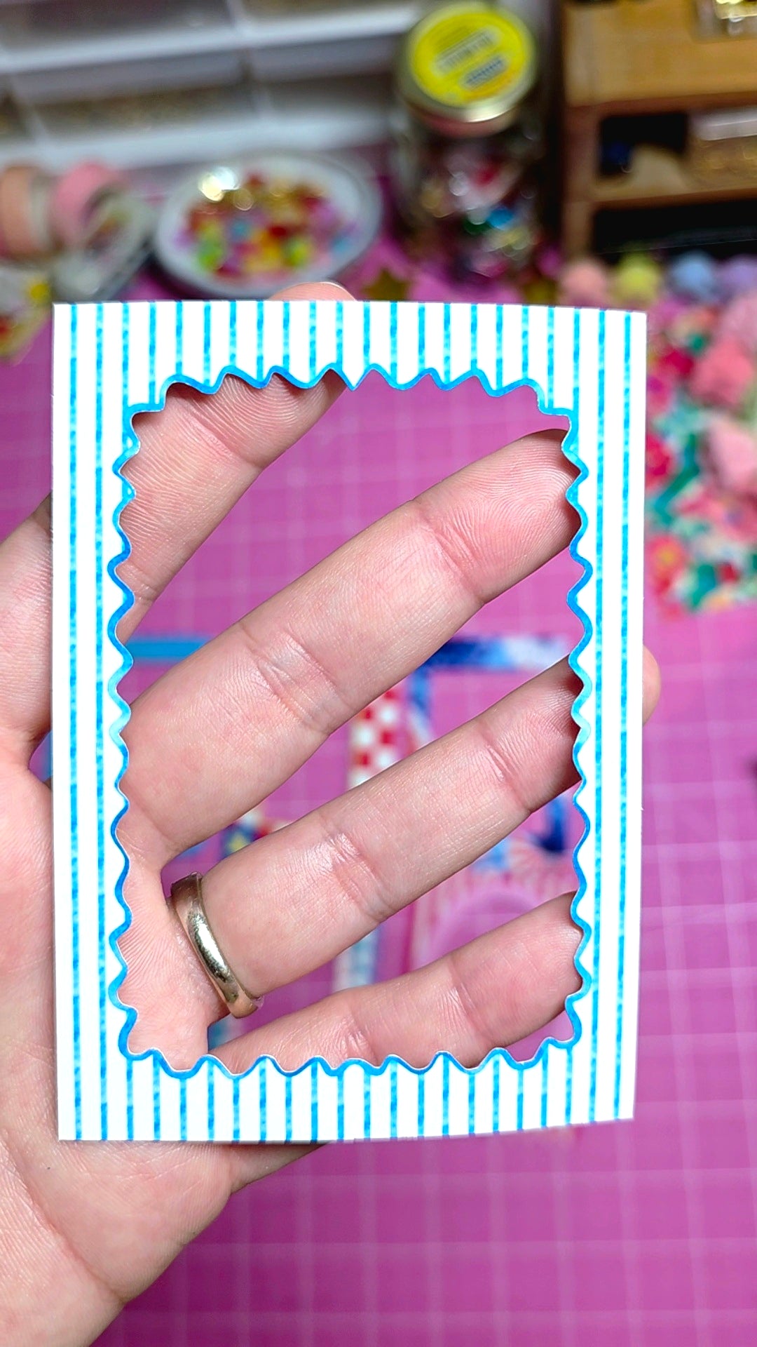 Hand with a ring on a pink surface with colorful objects in the background