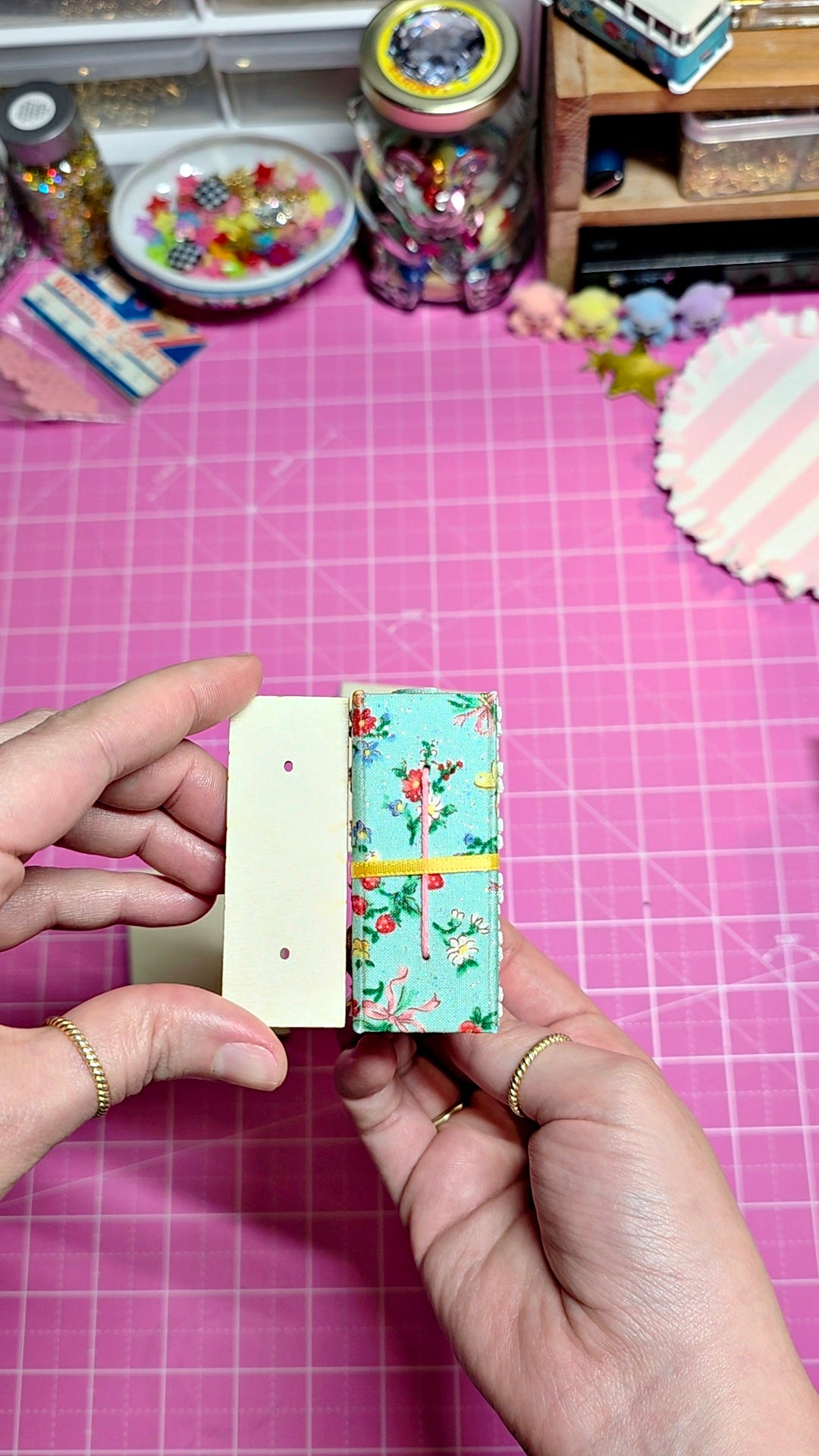 A pair of hands holding a small handmade paper craft box decorated with a floral pattern and a blank cream-colored card, set against a pink cutting mat background with craft supplies.