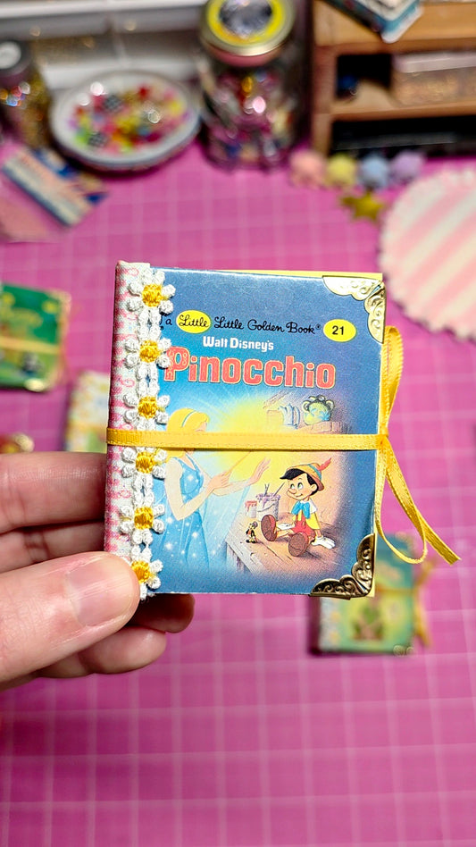A hand holding a miniature Pinocchio book journal with a yellow ribbon, set against a pink cutting mat background with other miniature items.