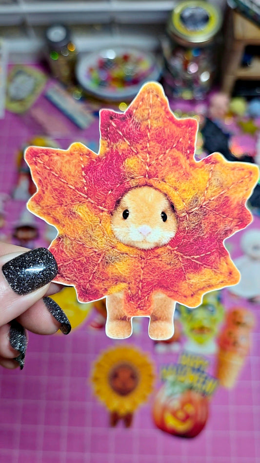 A hand with black glitter nail polish holds a die-cut sticker or patch shaped like a fall leaf with a small cartoon animal face in the center, set against a pink craft mat with other colorful items blurred in the background.