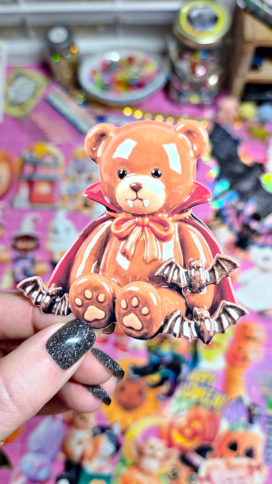 A hand with glittery black nails holds a small Halloween-themed teddy bear accessory featuring a vampire cape, bow, bat wings, and paw prints, set against a colorful, blurred background of stickers or cards.
