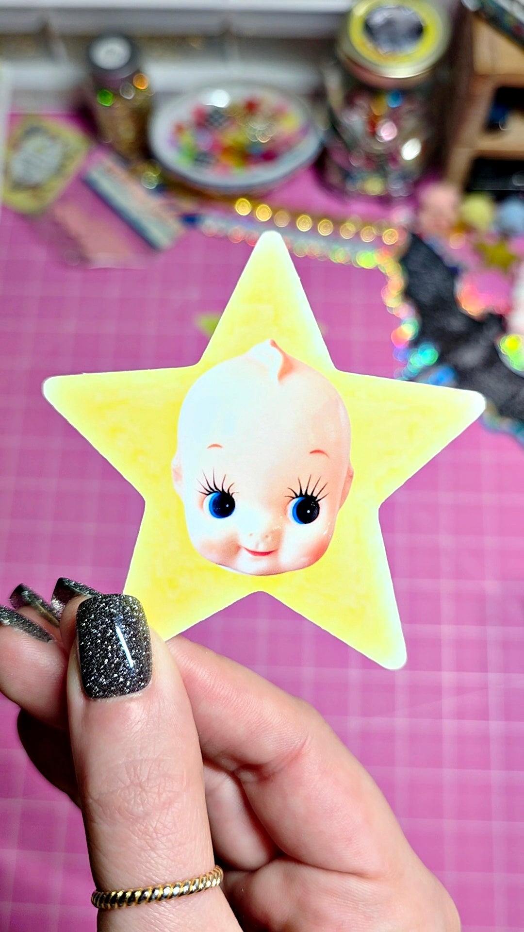 A hand with glittery black nail polish holds a yellow star-shaped sticker featuring a vintage-style baby face with large blue eyes and long eyelashes, against a pink craft mat background.