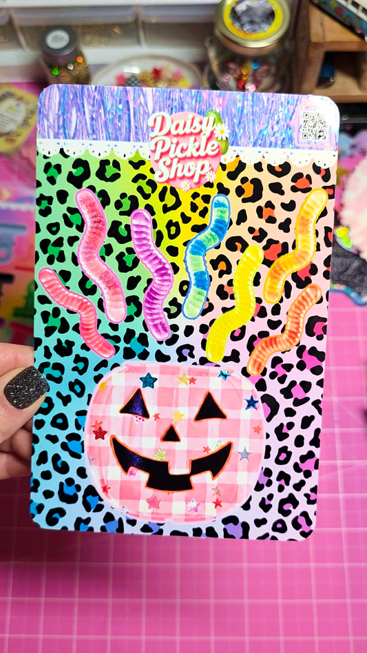 A hand with black glitter nail polish holds a decorative sticker sheet from Daisy Pickle Shop. The sheet features a Halloween theme with a pink gingham jack-o'-lantern, colorful wavy rainbow lines resembling teeth or hair, and a background of leopard print in pink, yellow, black, and blue.