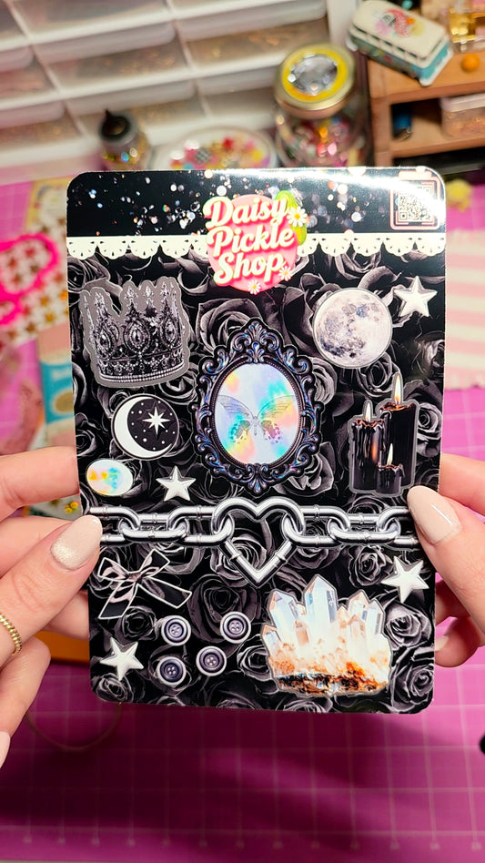 A person holds a decorative sticker sheet featuring a black gothic theme with stars, moons, crystals, and the 'Daisy Pickle Shop' logo. The background shows a craft room setting with a pink cutting mat.