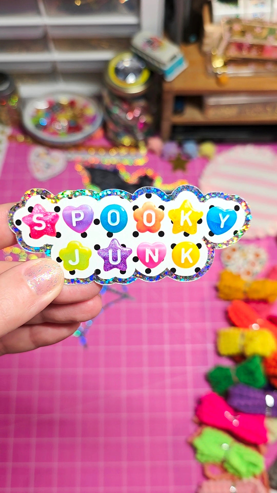 A hand holding a scalloped die-cut sticker that reads 'Spooky Junk' in colorful letters on a pink craft table with craft supplies in the background.