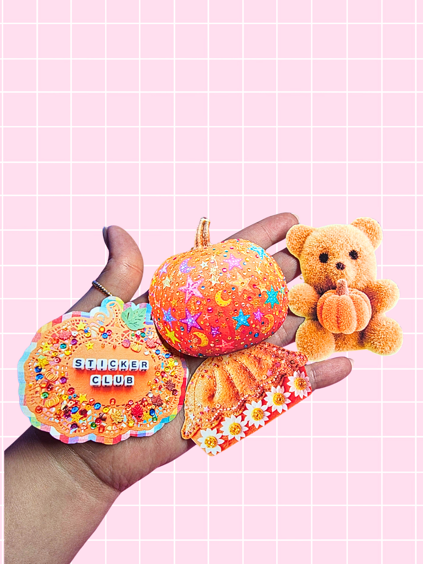 A hand holding three decorative autumn-themed stickers against a pink grid background: a pumpkin-shaped sticker with stars and moons, a round sticker reading 'STICKER CLUB' with colorful confetti, and a small plush teddy bear holding a pumpkin.