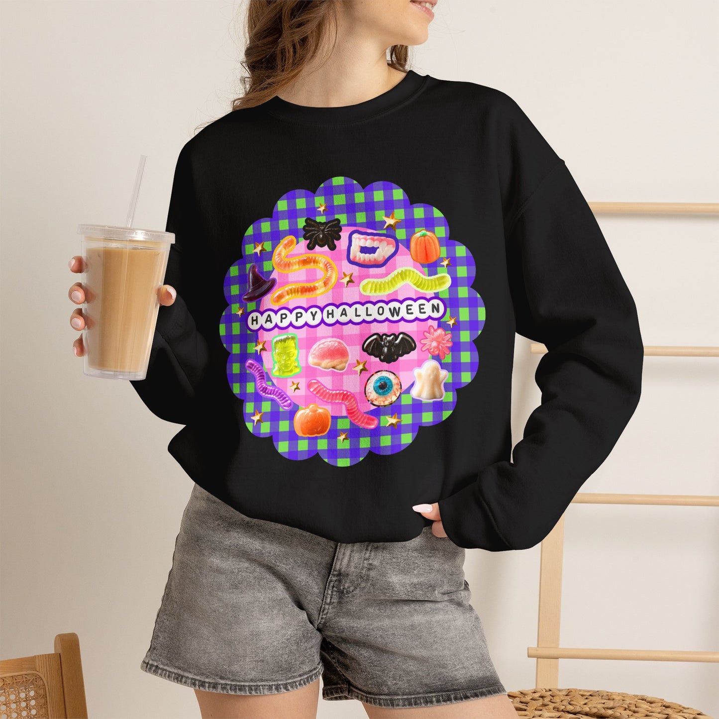 A woman wearing a black crewneck sweatshirt with a colorful 'Happy Halloween' graphic on the front, paired with gray denim shorts and holding an iced coffee.