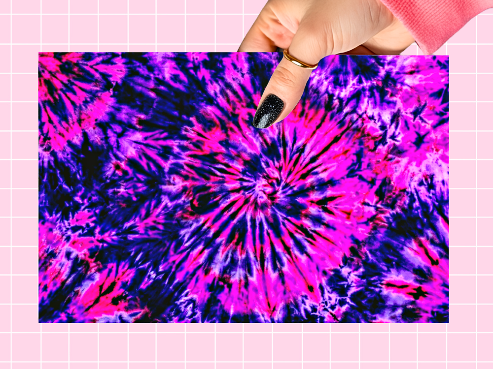 A hand with black glitter nail polish holds a square sample of a vibrant tie-dye pattern featuring a spiral design in hot pink, purple, and dark blue. The background is a light pink grid pattern.