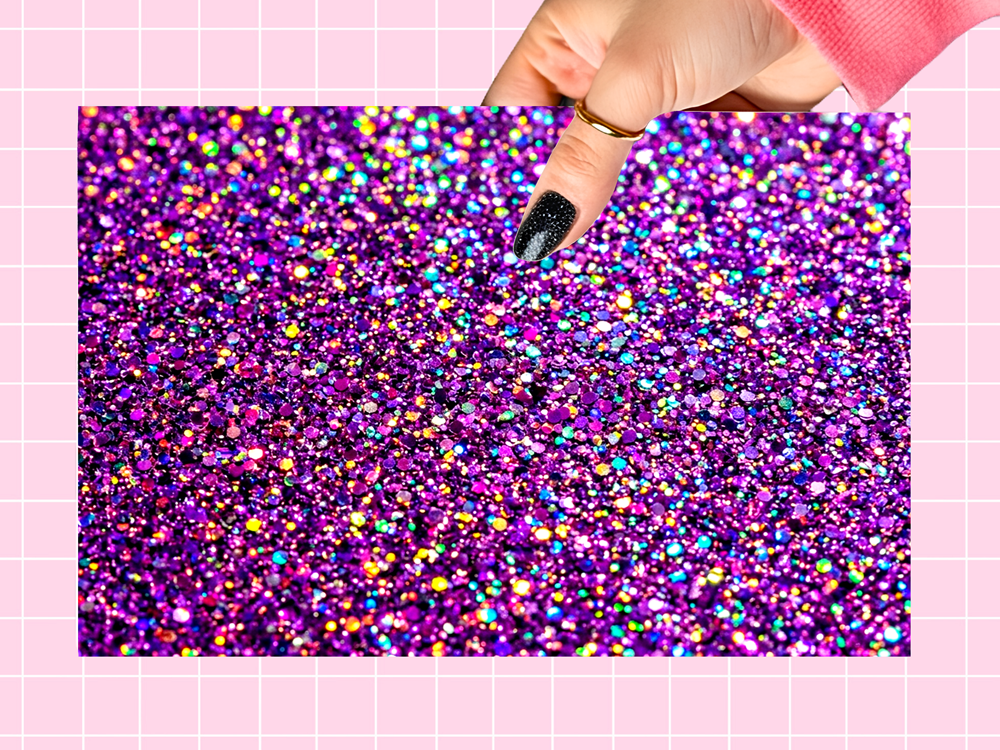 A hand with black glitter nail polish holds a rectangular sheet of vibrant purple holographic glitter against a pink grid background.
