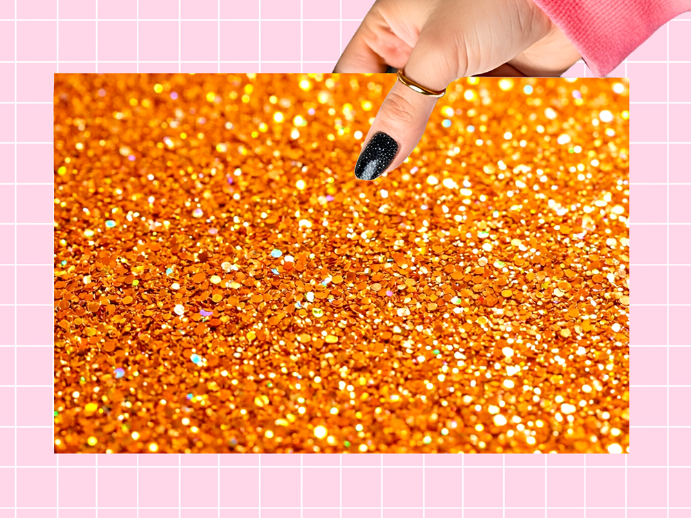 A close-up of sparkling orange glitter flakes, with a hand pointing at the texture. The image is framed by a light pink grid background.