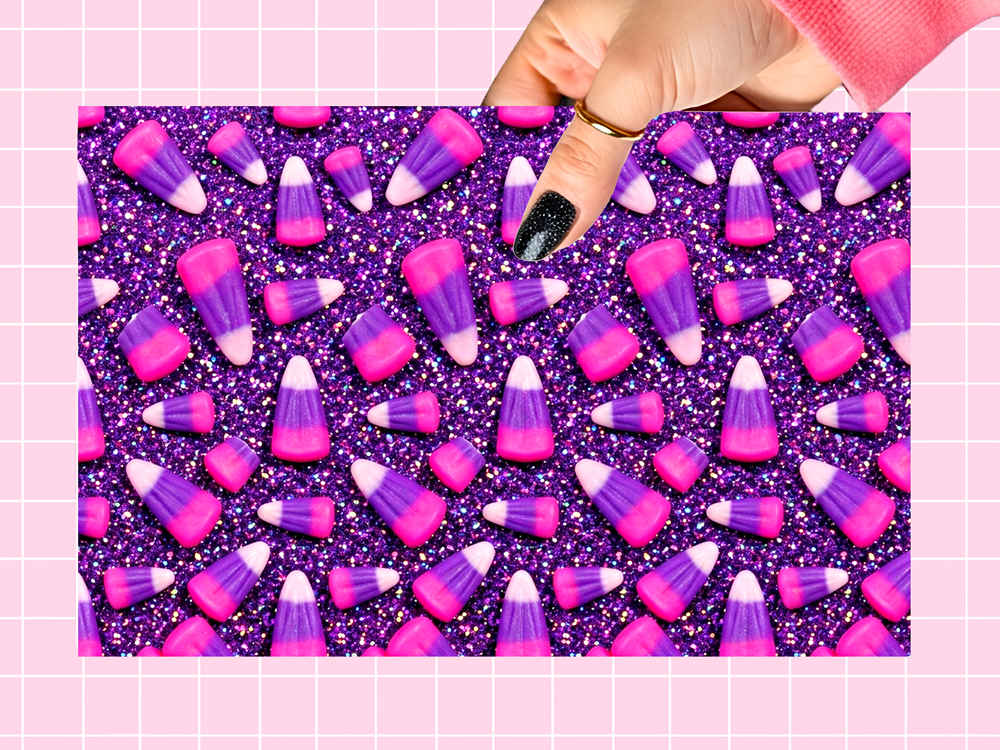 A rectangular sheet or card featuring a repeating candy corn pattern on a glittery purple background, held by a hand with black glitter nail polish against a pink grid backdrop.