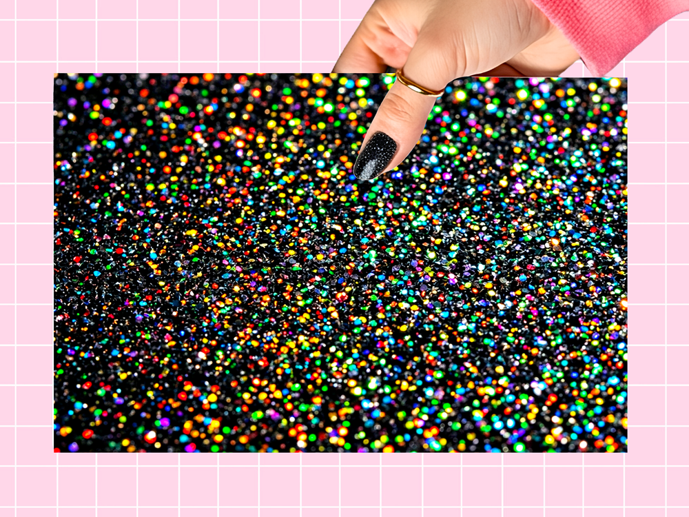 A rectangular black glitter mat with dense holographic rainbow glitter, held by a hand with black glitter nail polish against a pink grid background.