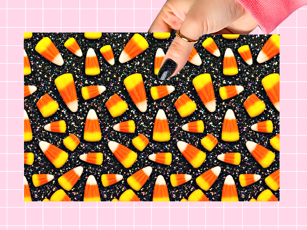 A rectangular sheet or card featuring a repeating candy corn pattern on a black glittery background, held by a hand with black glitter nail polish against a pink grid backdrop.