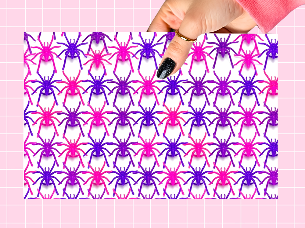 A hand with black glitter nail polish holds a rectangular sheet of colorful plastic toy spiders in pink and purple, set against a pink grid background.