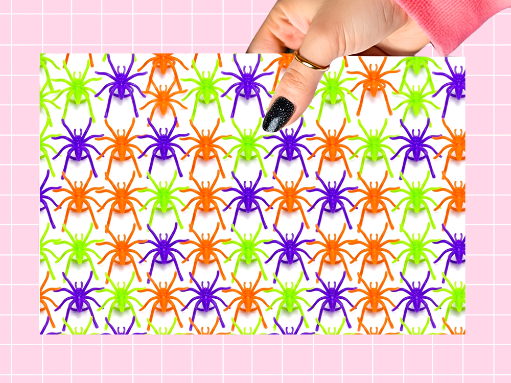 A hand with black glitter nail polish holds a rectangular card or sheet featuring a repeating pattern of colorful plastic toy spiders in purple, orange, and neon green on a white background, set against a pink grid backdrop.