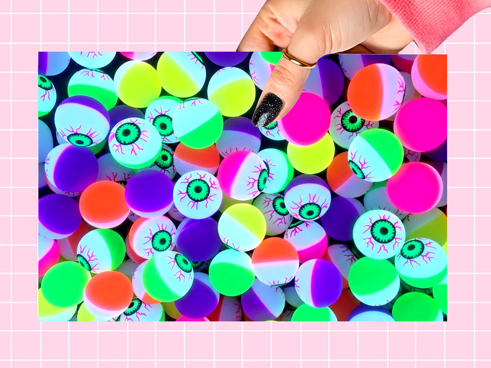 A hand with black glitter nail polish points toward a pile of neon-colored gummy candies shaped like eyeballs, set against a pink grid background.