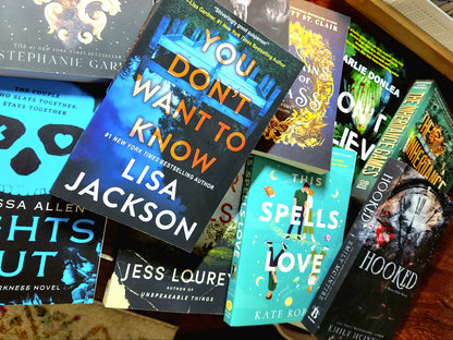 A collection of various items including a book, a hot chocolate, magnetic bookmarks, a headband, a beanie, socks, a highlighter, stickers, earrings, a face mask, and a bookmark, all arranged on a wooden surface.