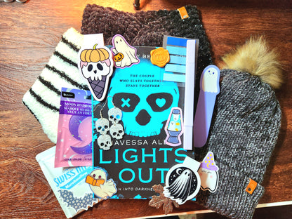 A collection of various items including a book, a hot chocolate, magnetic bookmarks, a headband, a beanie, socks, a highlighter, stickers, earrings, a face mask, and a bookmark, all arranged on a wooden surface.