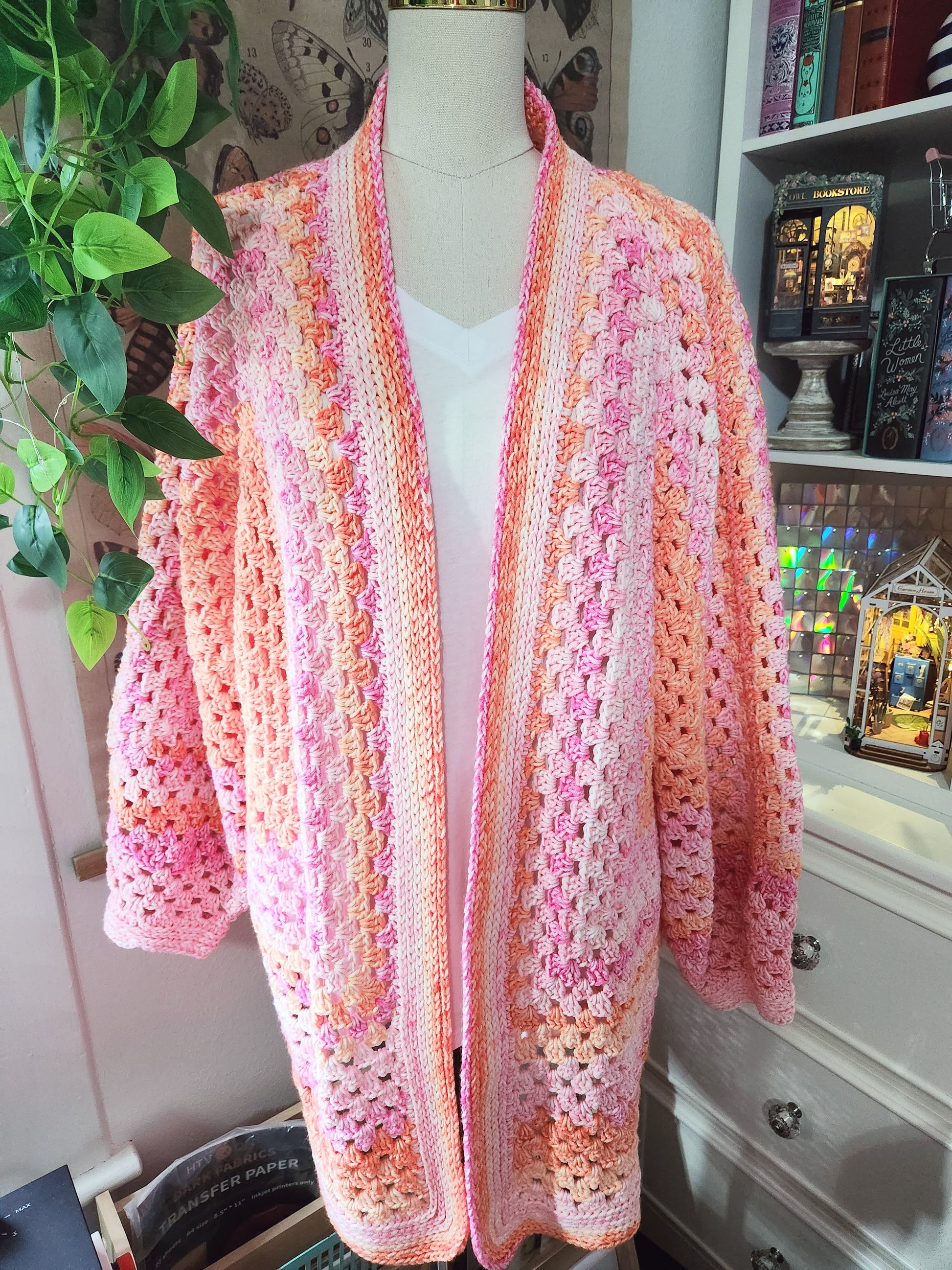 Mannequin wearing a pink and orange crocheted cardigan in a room with books and decor.