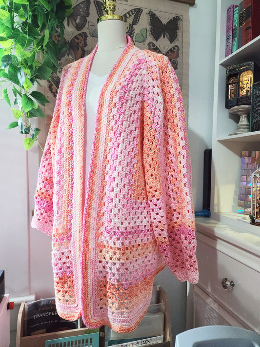 Crocheted cardigan with a gradient of pink, orange, and white on a mannequin against a home interior background.
