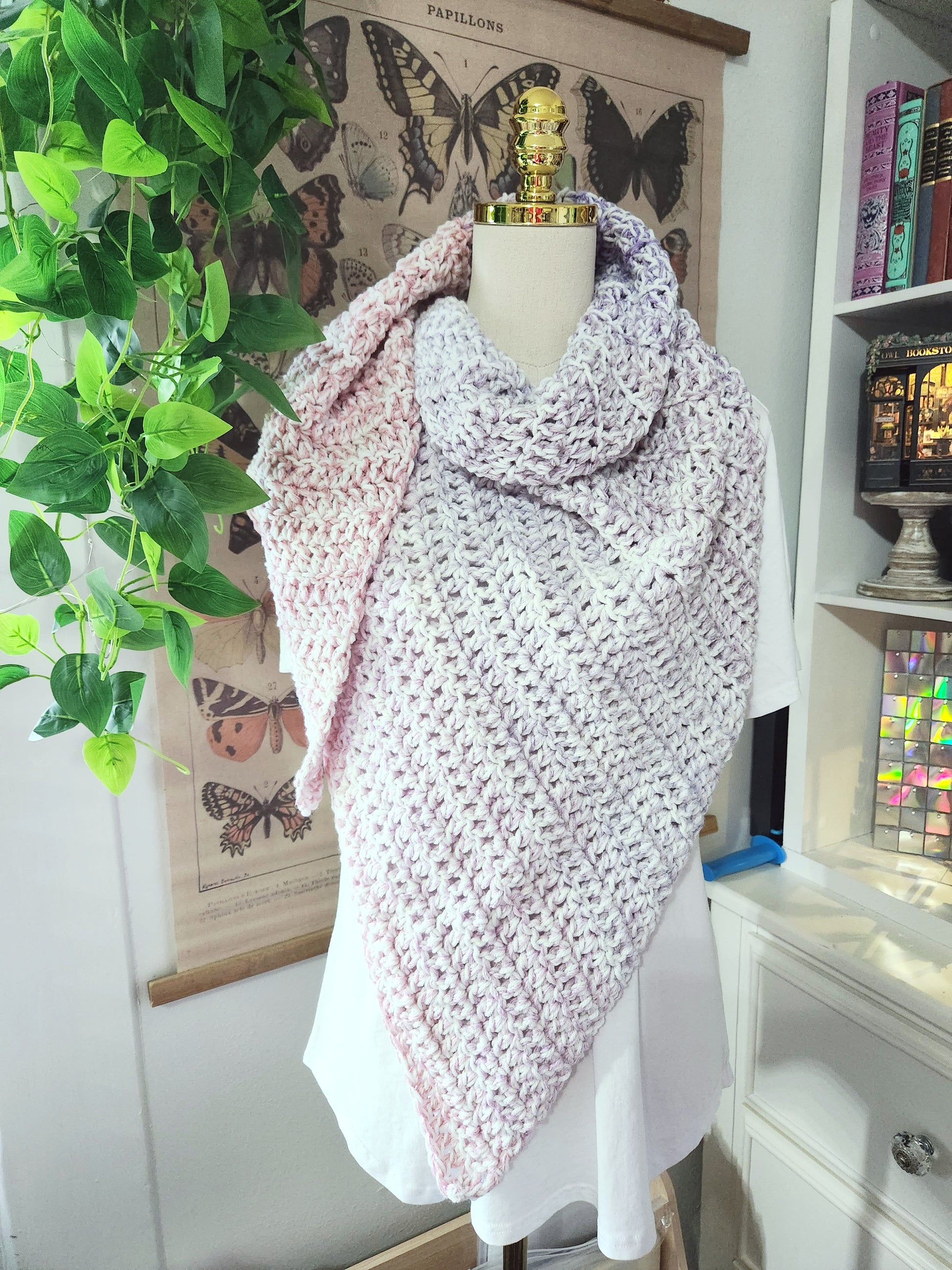 Mannequin wearing a pink knitted shawl in a room with plants and books.
