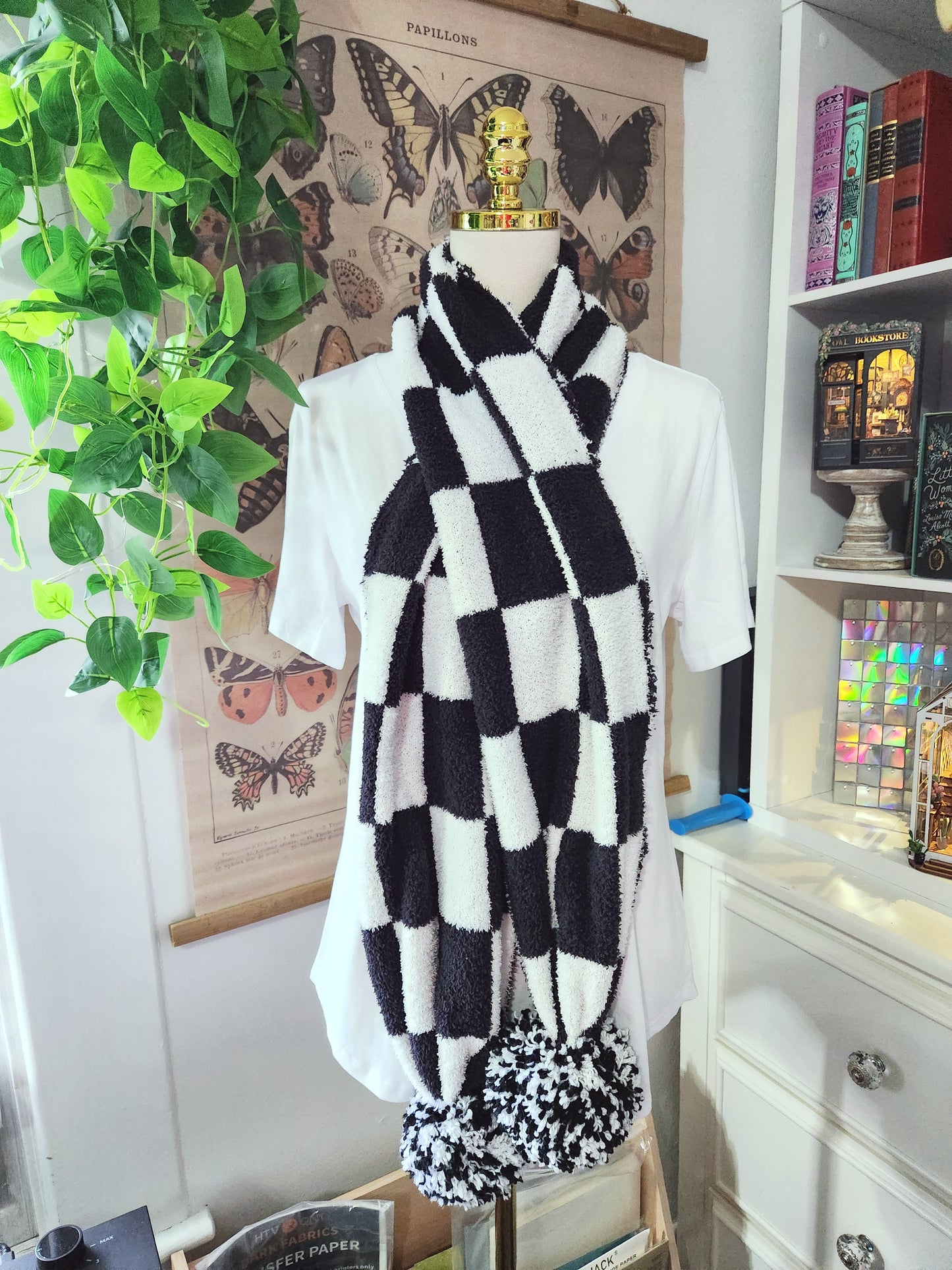 Black and white checkered scarf on a mannequin against a decorative wall with books and plants.