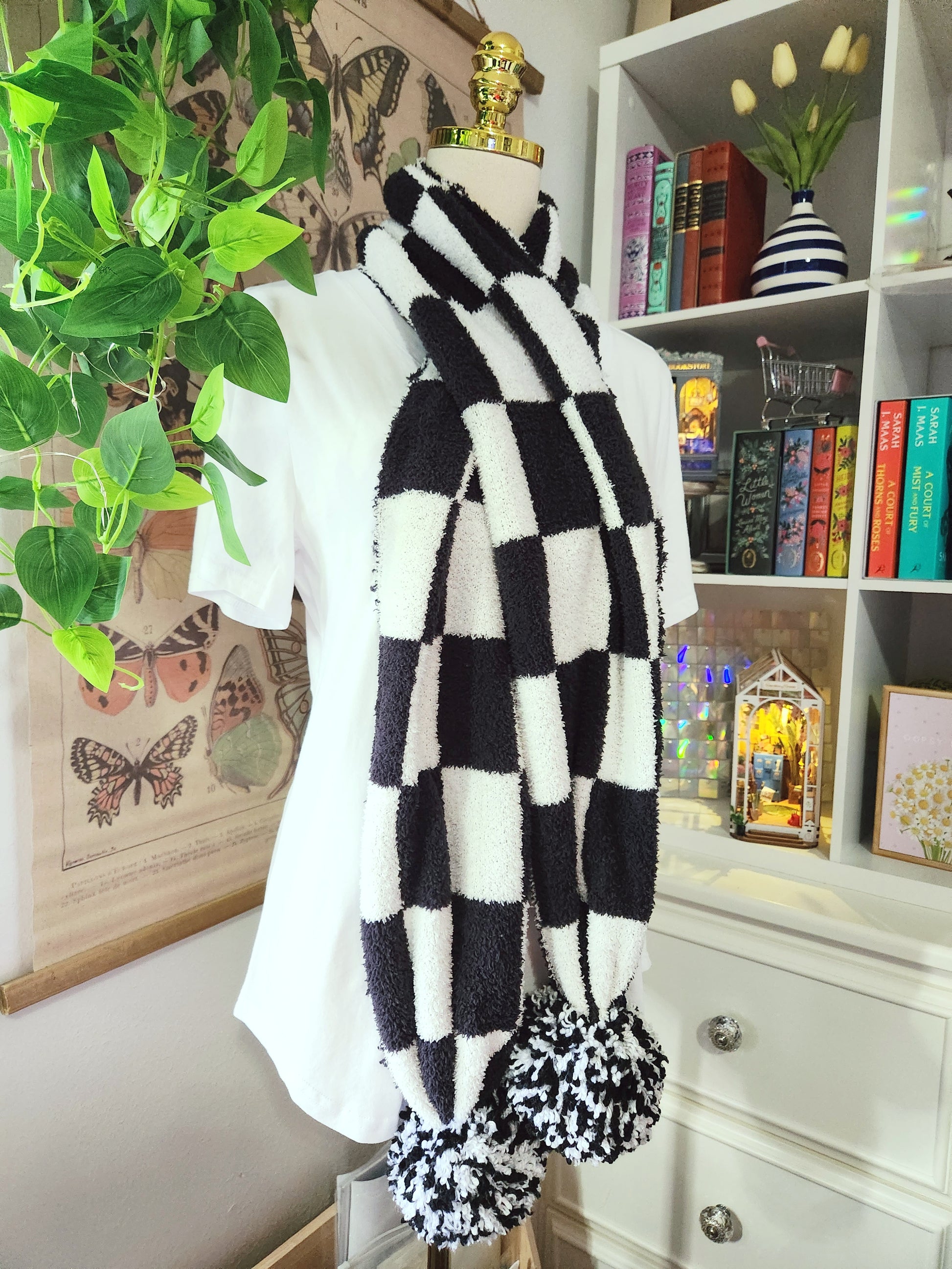 Black and white checkered scarf with pom-poms on a mannequin against a bookshelf background