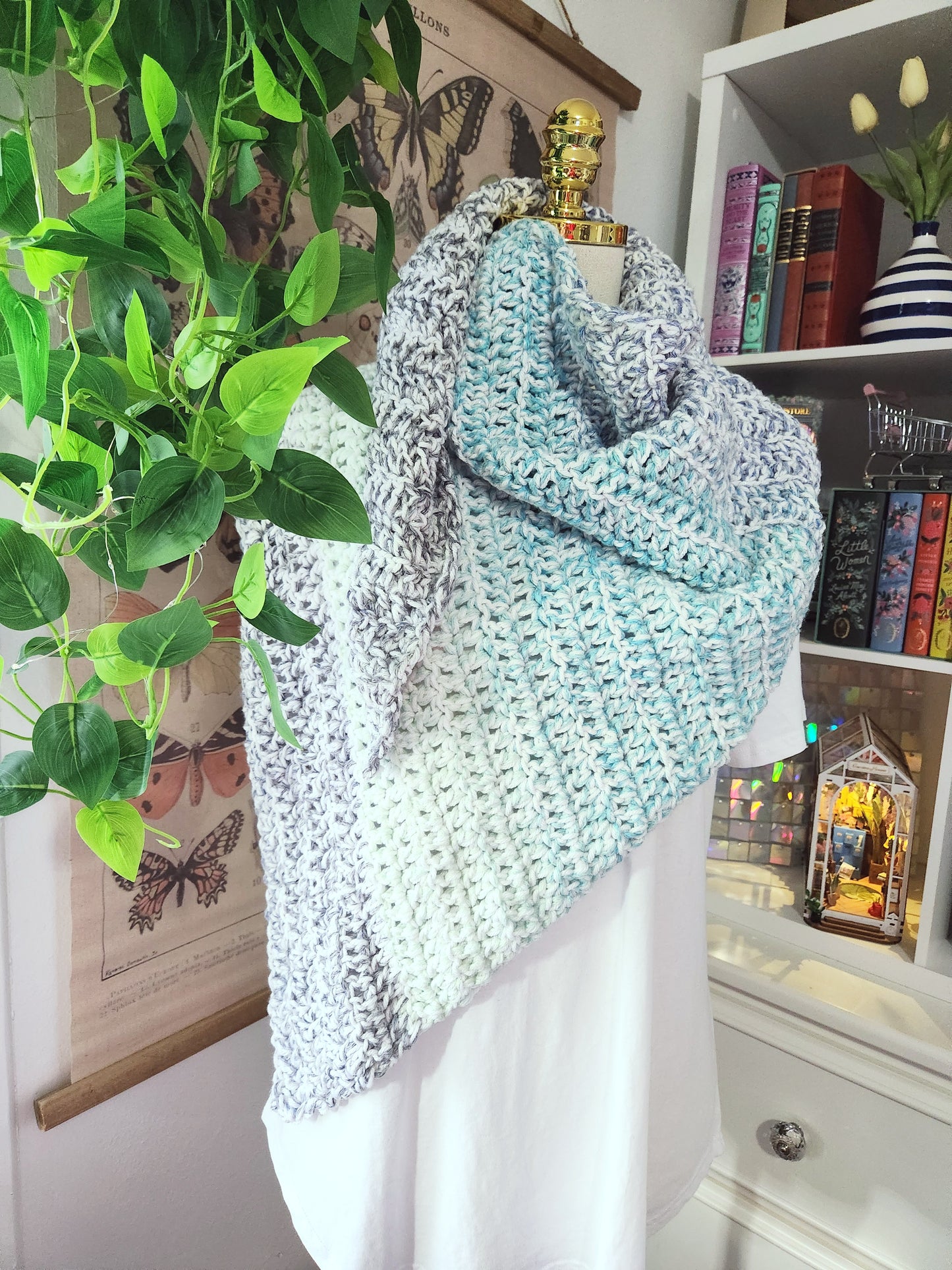 Knitted shawl on a mannequin with a decorative background including books and plants.