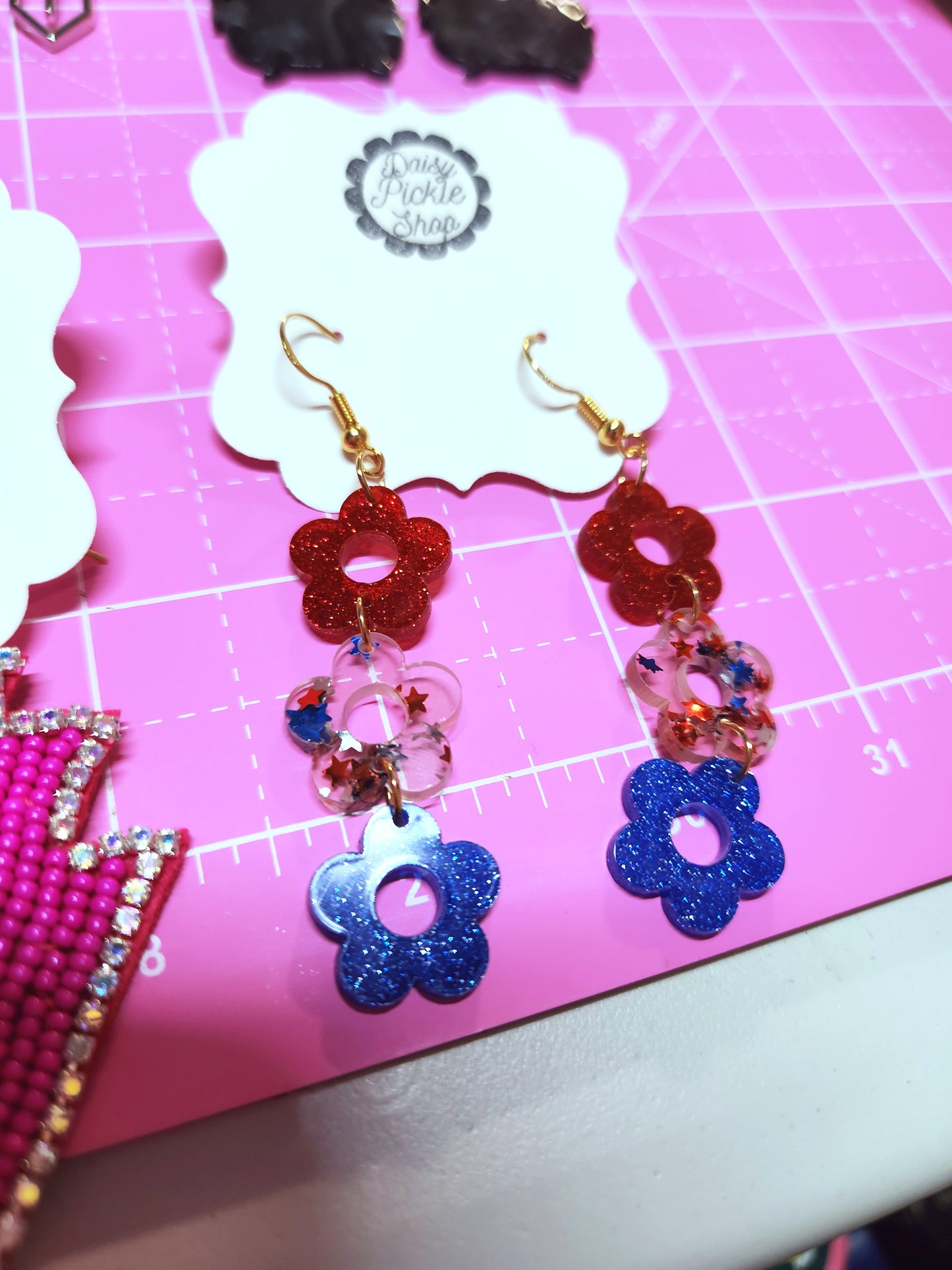 Colorful flower-shaped earrings on a pink cutting mat with a visible brand label.