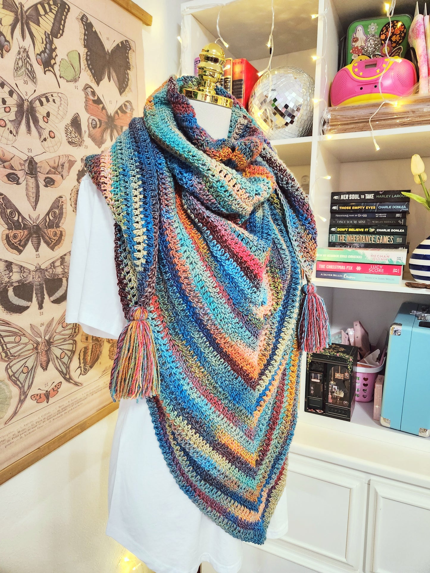 Colorful crocheted shawl on a mannequin against a decorative wall with butterfly illustrations.