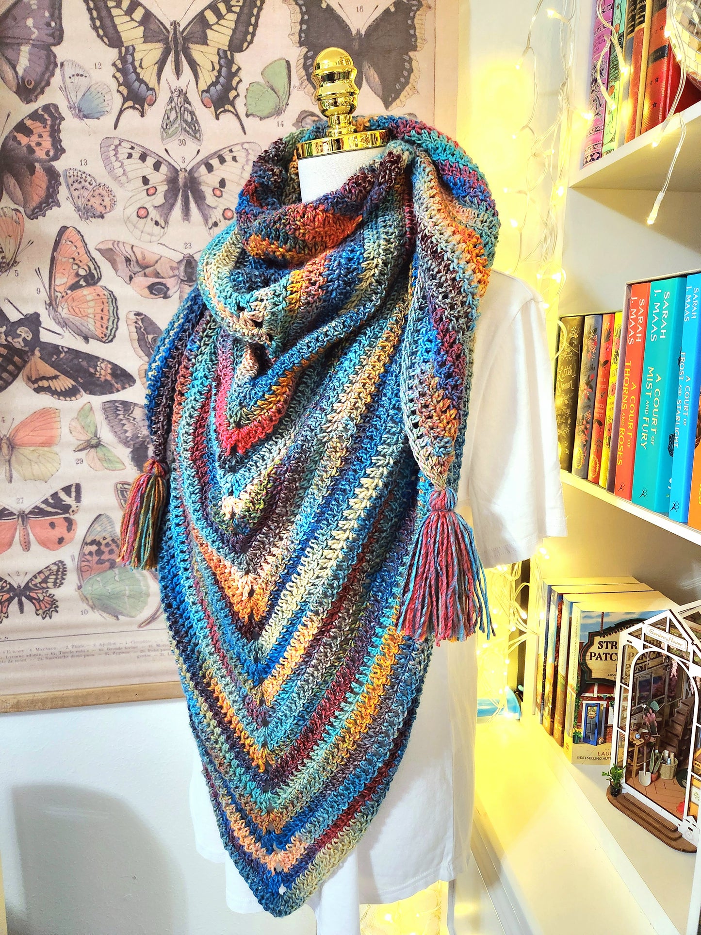 Colorful crocheted shawl on a mannequin against a background with books and butterfly decorations.
