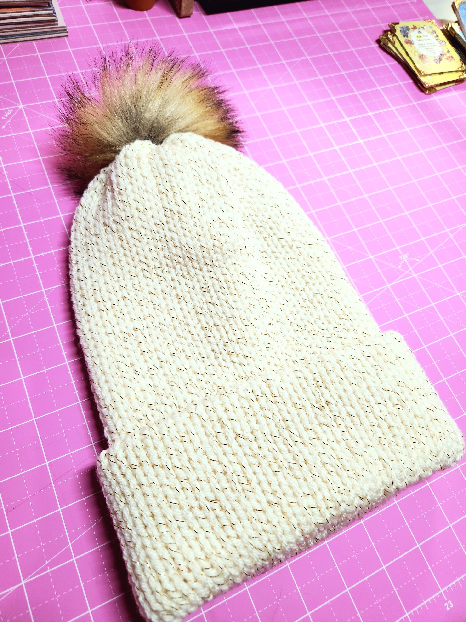 Knitted white heart-shaped item with a fluffy brown pom-pom on a pink cutting mat.