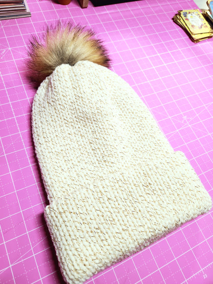 Knitted white heart-shaped item with a fluffy brown pom-pom on a pink cutting mat.