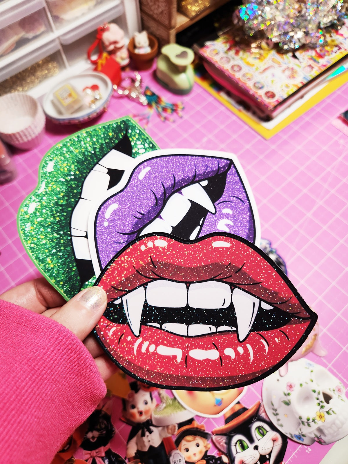 Colorful glittery lips patch held by a hand on a pink surface with toys in the background