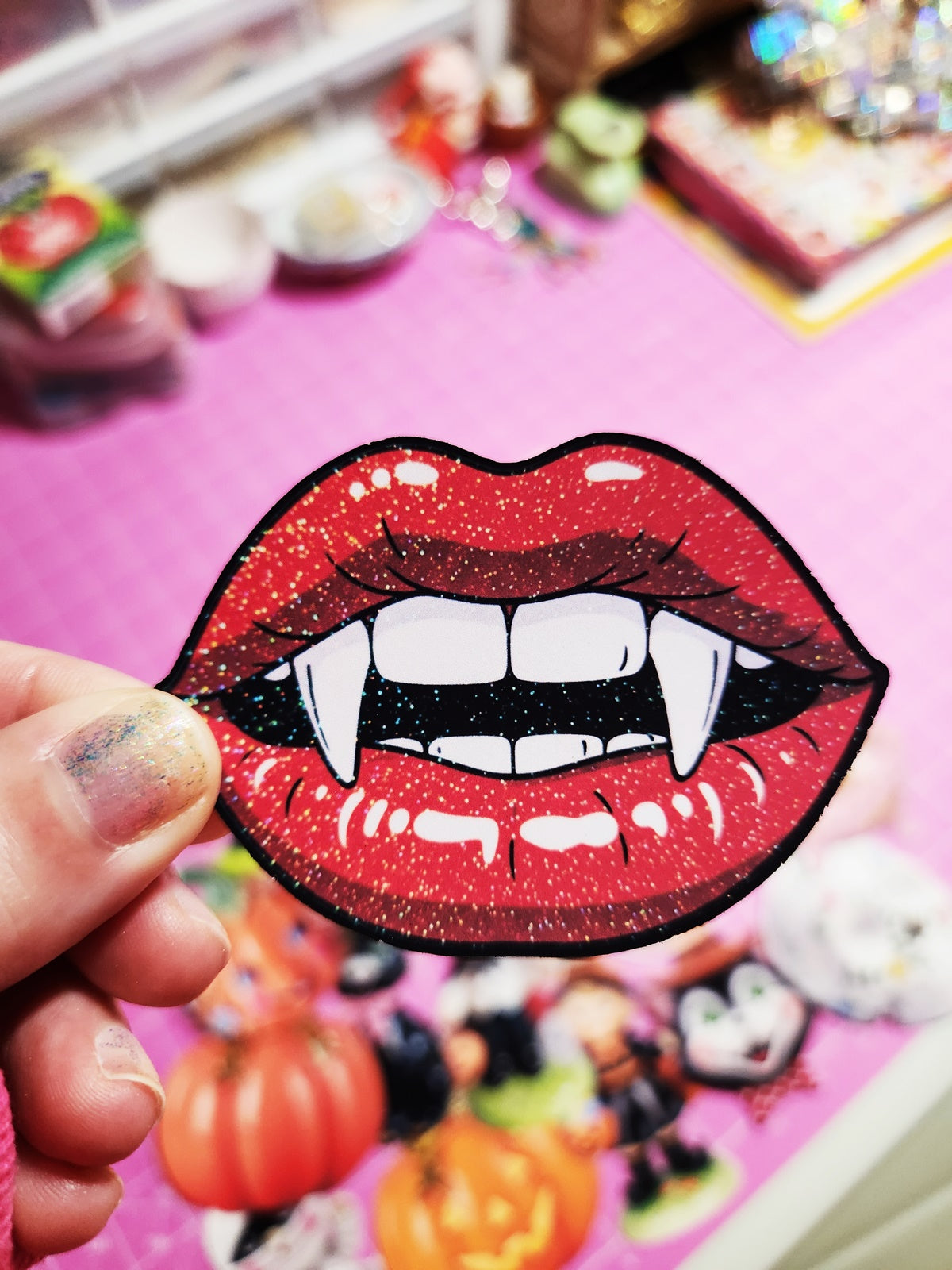 Red glittery lip patch with vampire teeth held by a hand on a colorful Halloween-themed background.