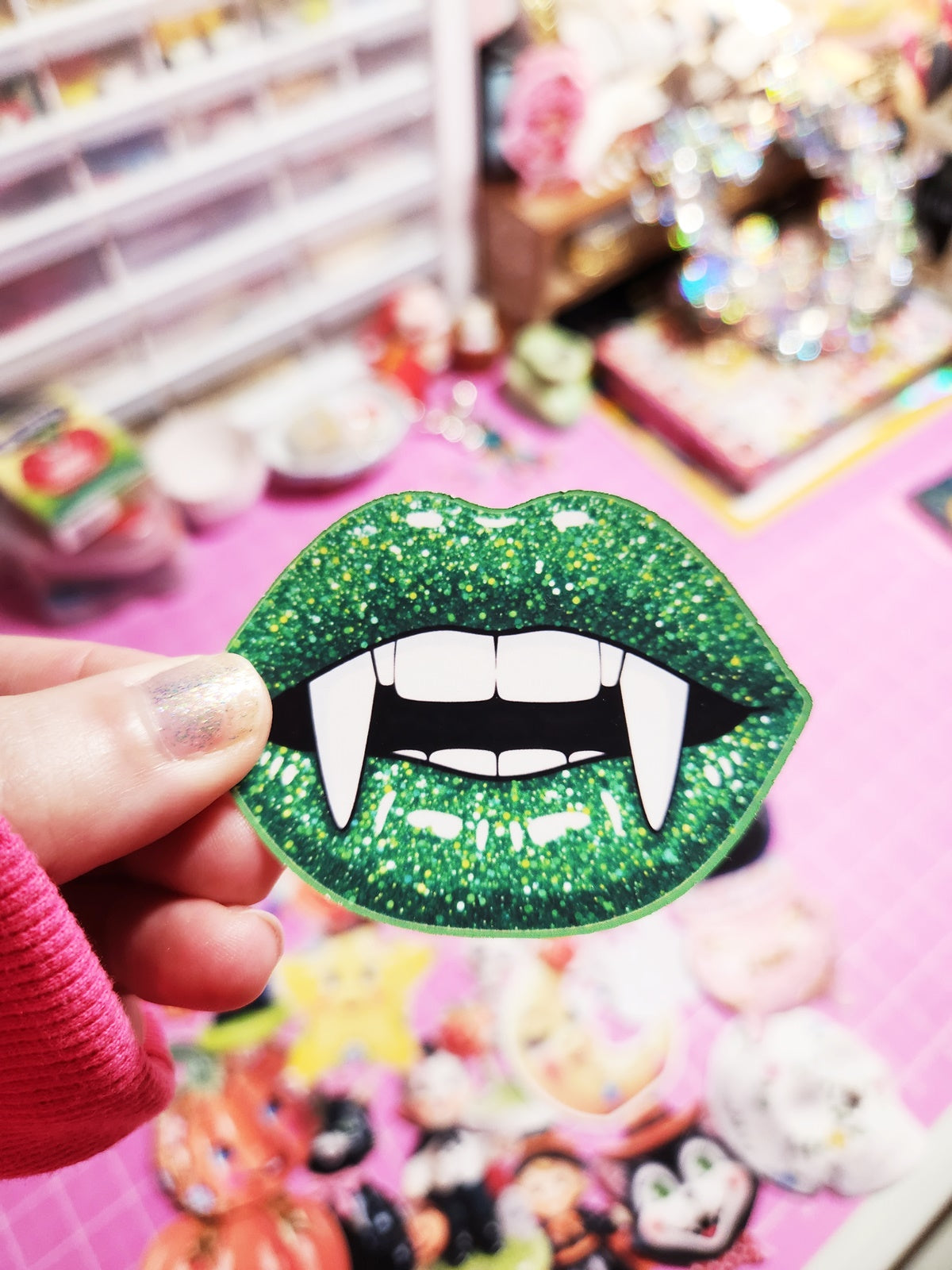Green glittery vampire lips sticker held by a hand against a colorful, blurred background