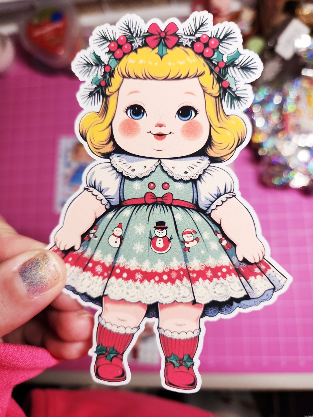 Illustration of a girl in a festive dress with snowmen and red bows, held against a pink background.