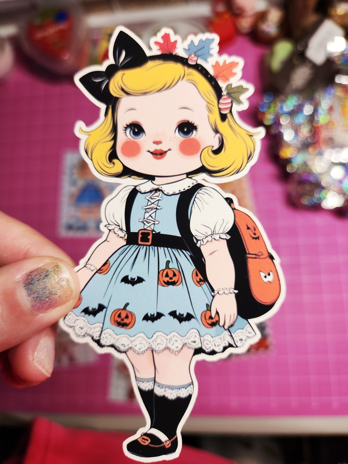 Sticker of a girl with blonde hair and a blue dress with Halloween-themed decorations on a pink background.