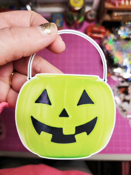 Green Halloween candy bucket with a face design held by a hand against a colorful background.