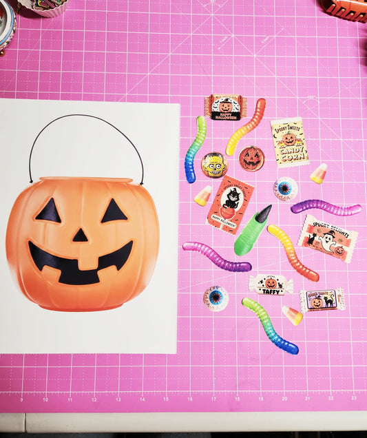Halloween-themed candy and decorations on a pink cutting mat with a pumpkin bucket template.
